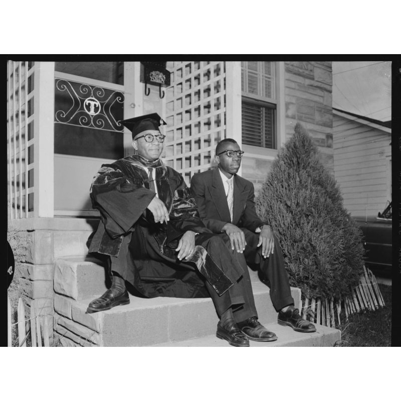 Two men sitting on the front steps of a house, one in graduation regalia.