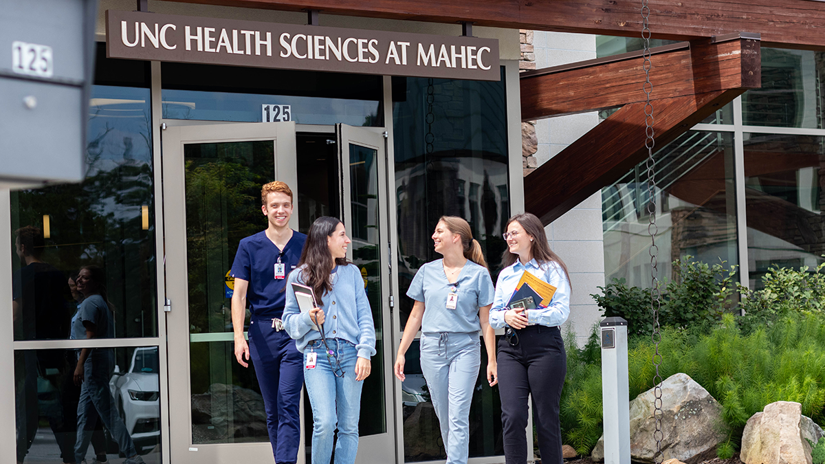 Four medical students wearing scrubs and holding books exit building at the School of Medicine's Asheville campus. Sign above front door reads 